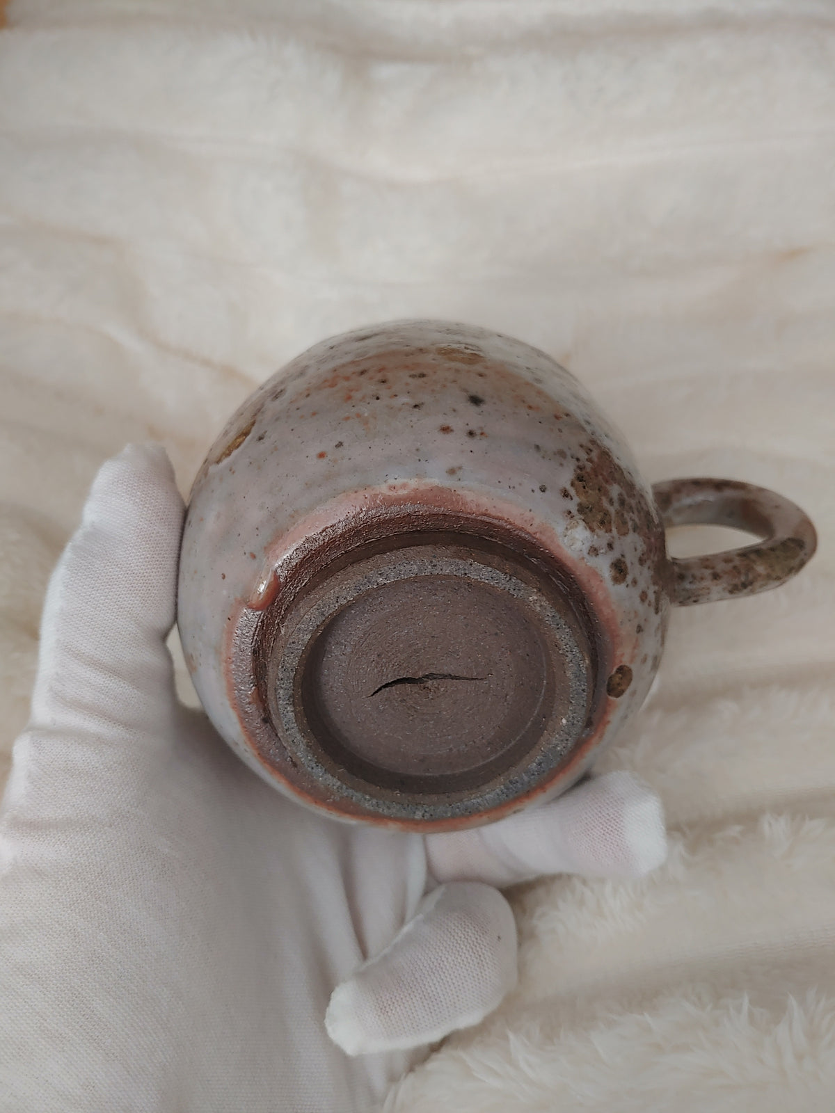 A handmade ceramic mug with a handle, held by a white gloved hand against a soft, textured cream fabric background. The mug features a speckled, earthy glaze with brown, gray, and muted pinkish-brown tones.