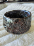 A handmade, rustic ceramic ring bowl with a textured, earthy glaze in dark brown, black, and subtle greenish tones, resting on a white faux fur surface.
