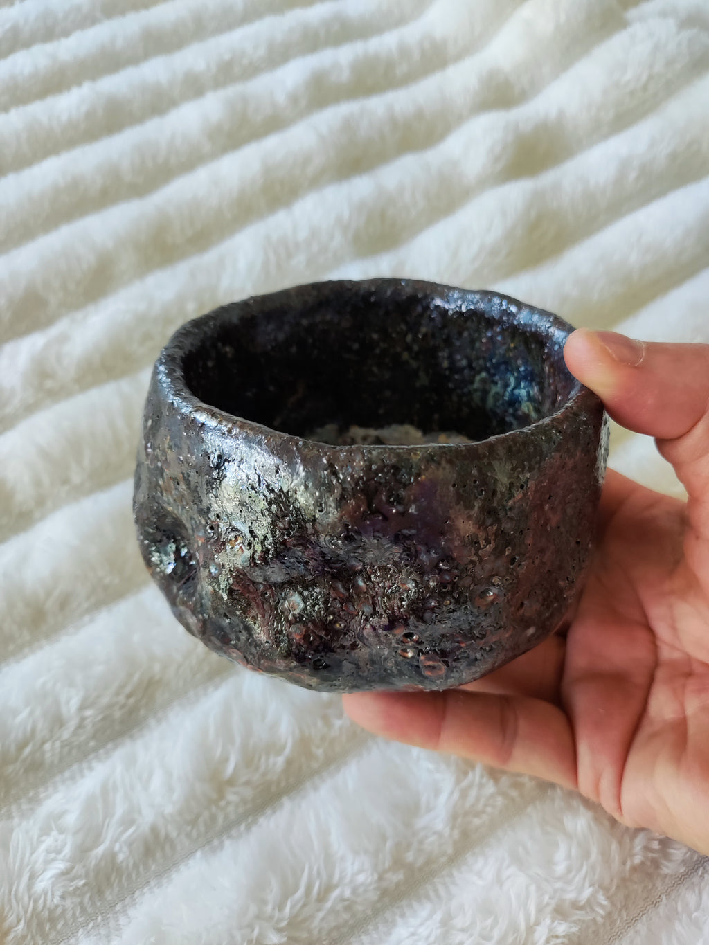 A handmade ceramic bowl with a dark, textured glaze and metallic copper-brown speckles, held in a person's hand against a white textured blanket background.