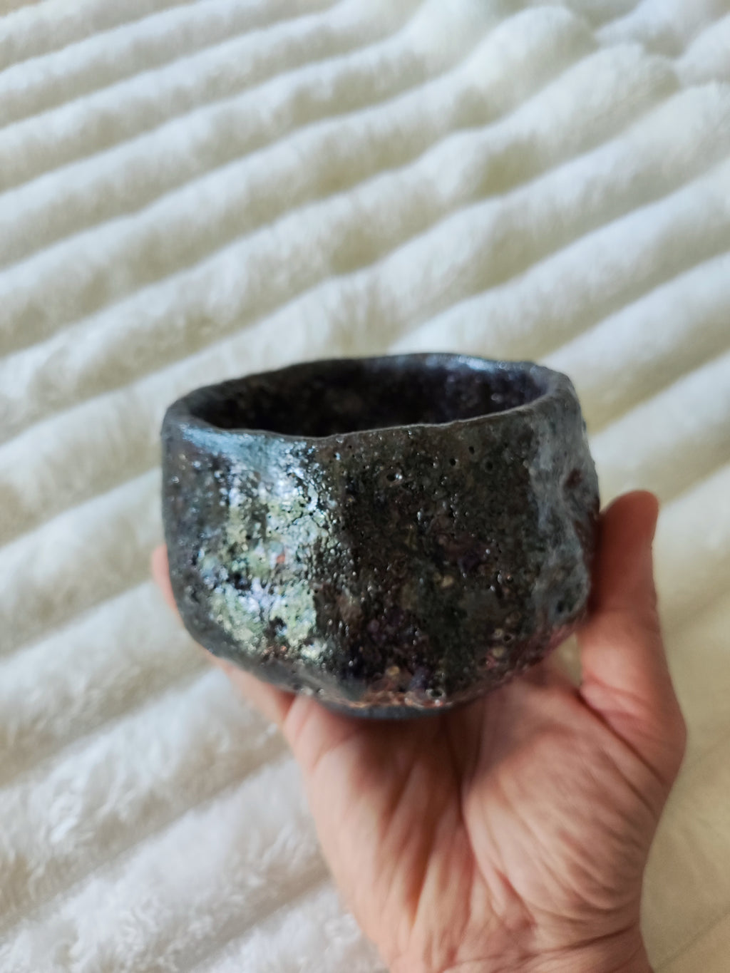 A handmade dark ceramic bowl with a textured, speckled finish, held in a person's hand against a white ribbed fabric background.