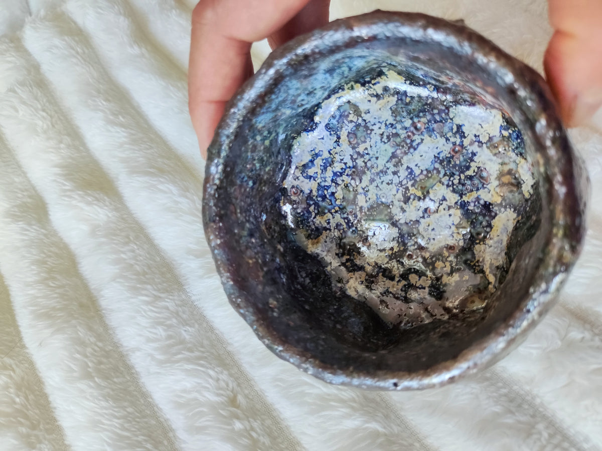 A handmade ceramic bowl with a rustic, textured finish and a speckled glaze of blue, gray, and white, held by hands over a white faux fur background.