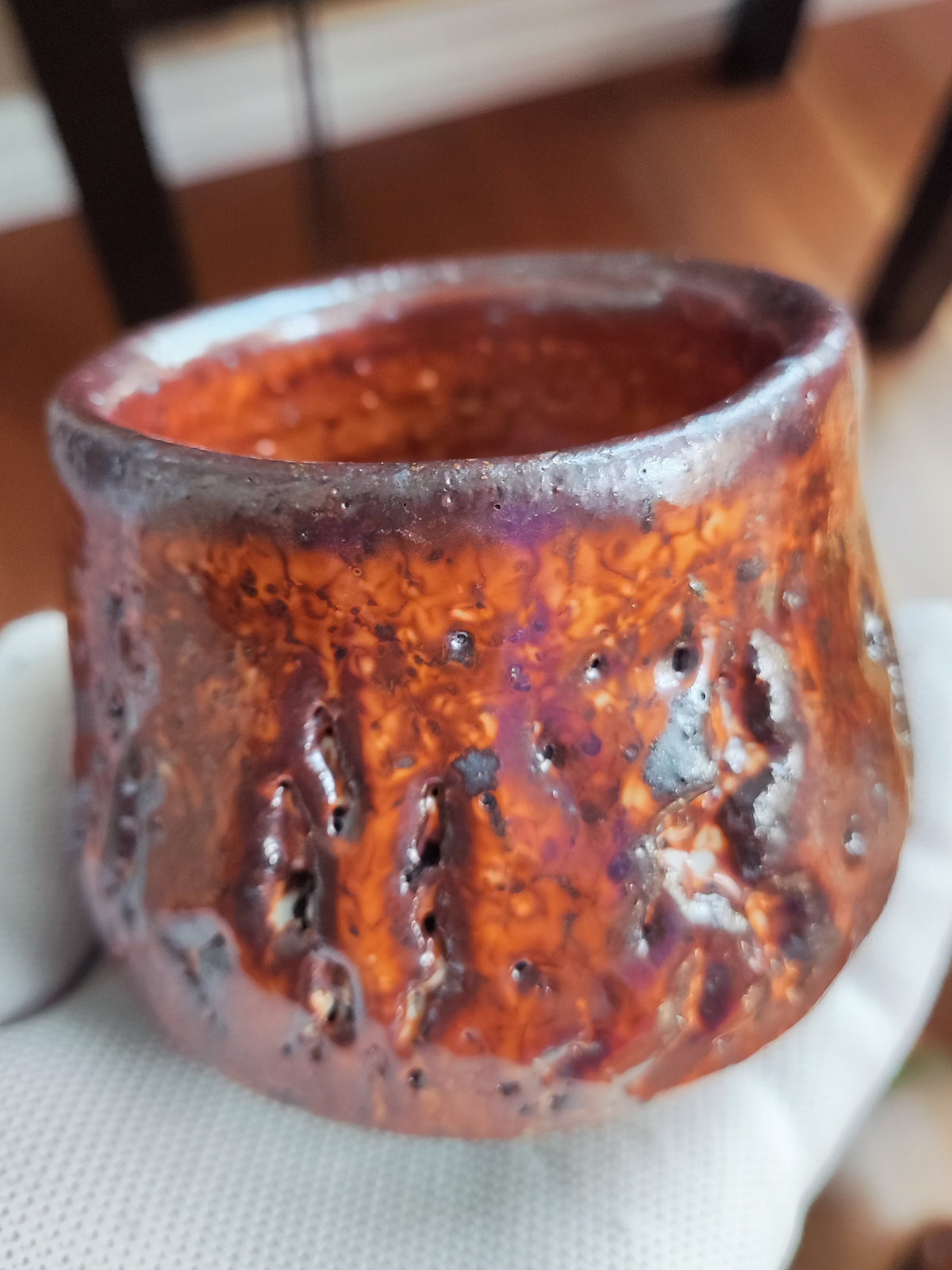 A handmade ceramic cup with a textured, bubbly orange-brown glaze, held in a white mesh glove. The background shows a blurry indoor setting with wooden floors and furniture.